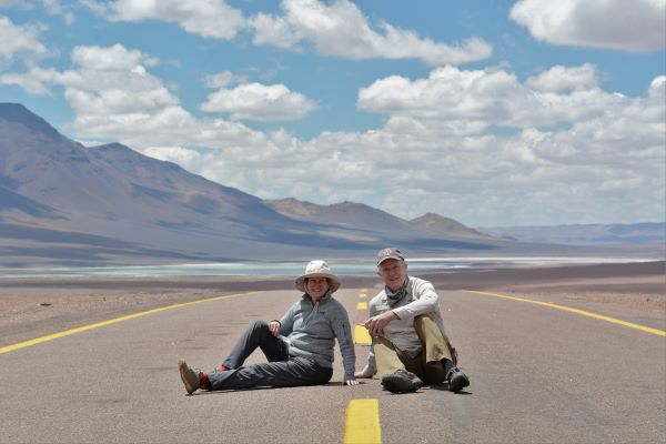 A man and a woman sit in the middle of an empty highway with dry, desert mountains and a bright blue, cloudy sky reflecting off the valley floor behind.