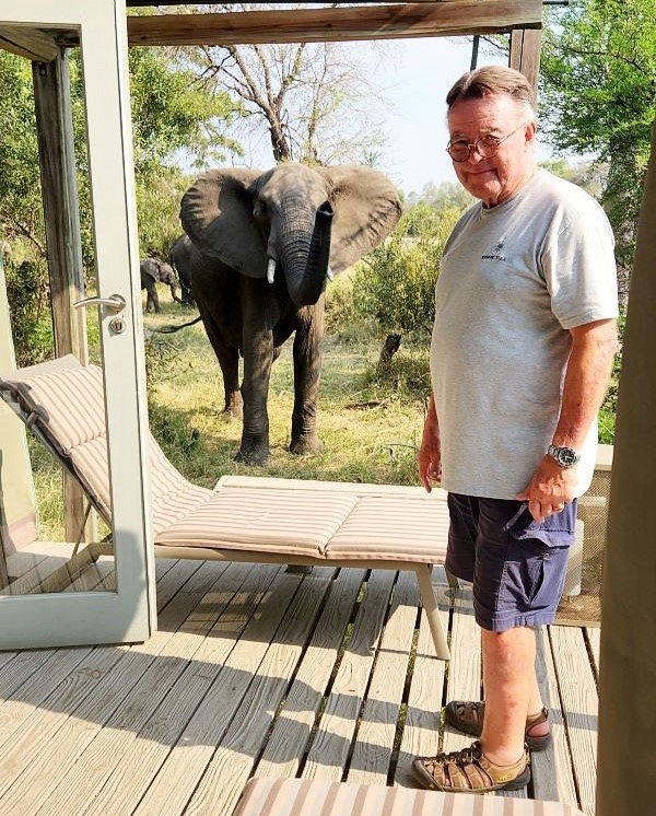 A man stands next to a chaise lounge and an open door on his veranda with a large elephant standing just beyond, looking straight at him and the camera.