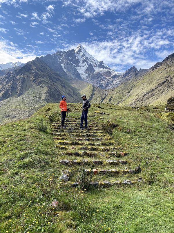 Two hikers on a green grassy trail staircase with a large mountain peak in the background.